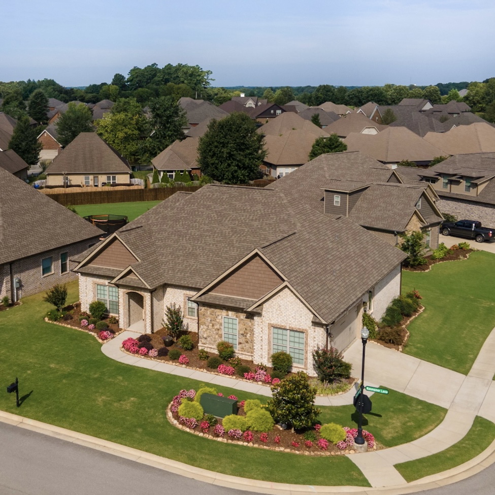 Aerial Perspective of Elegant Corner Lot Home "Drone view of a house on a corner lot with off-white brick and stacked stone siding, brown roof, and well-manicured yard with flowering shrubs in Huntsville, AL.